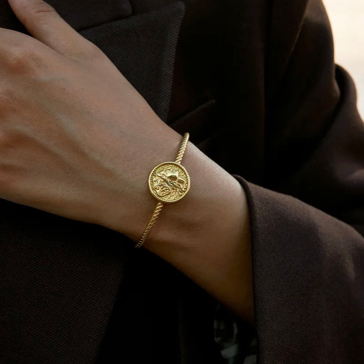 Gold bracelet with a coin design worn on a wrist against a dark background